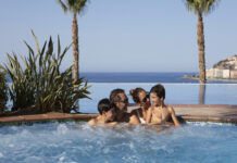 Una pareja con dos hijos en una piscina panorámica mirándose entre sí, con palmeras y el mar de fondo