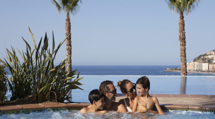 Una pareja con dos hijos en una piscina panorámica mirándose entre sí, con palmeras y el mar de fondo