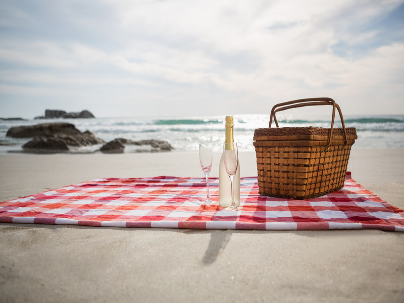 Two glasses, champagne bottle and picnic basket on beach blanket at tropical beach
