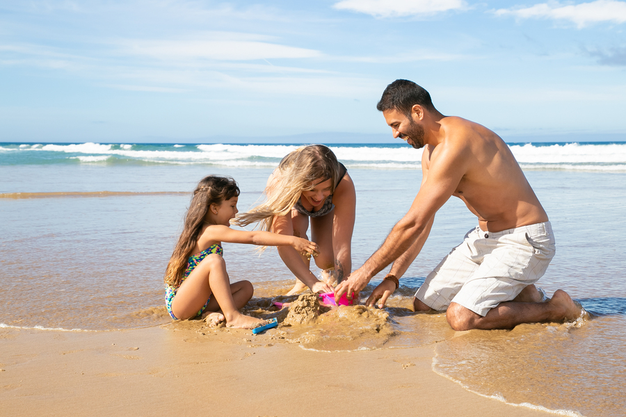 Joyful mom, dad and little daughter enjoying vacation at sea together, playing with daughters sand toys, building sandcastle. Family summer holidays concept